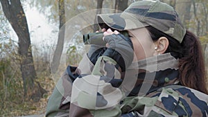 Beautiful woman in army camouflage looks through binoculars away on the river in the fog on a cold morning