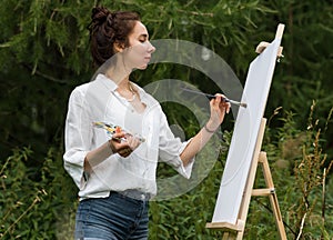 Beautiful woman artist, in summer in park draws a picture, a palette with paints, a brush and a white canvas painting