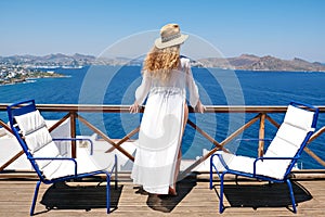 Beautiful woman from back in swimsuit white dress and straw hat on white terrace balcony of house or hotel with Sea View