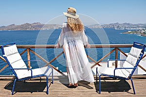 Beautiful woman from back in swimsuit white dress and straw hat on white terrace balcony of house or hotel with Sea View