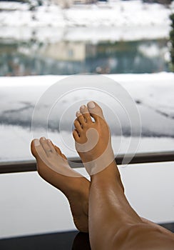 Beautiful woman bare feet up on the table in warm classy interior with large glass window and luxury view of the snowy landscape