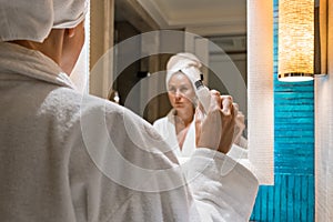 Beautiful woman in bathrobe holding a jar of cream applying face skin moisturizer, standing in front of bathroom mirror