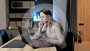 Beautiful woman in beige sweater works late at home at her desk at laptop. Concept freelance