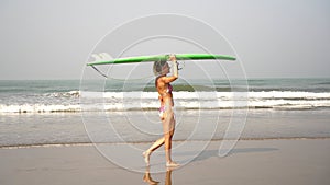A beautiful woman in a bikini walks along the beach against the sea with a surfboard