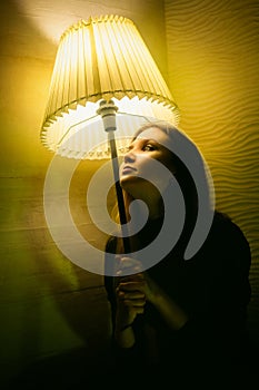 A beautiful woman in black clothes holding a floor lamp and gazing at a glowing lamp on a dark evening. Self-reflection