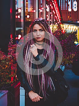 Beautiful woman in black clothes with long pink dreadlocks at night against the background of multicolored lights