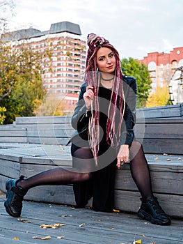 Beautiful woman in black clothes with long pink dreadlocks sits on wooden steps and looks at the camera