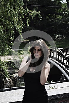 Beautiful woman in black dress is touching her hair and smiles in the park near the bridge and trees