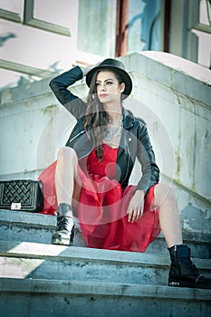 Beautiful woman with black hat, red dress and boots posing sitting on stairs. Young brunette spending time during autumn