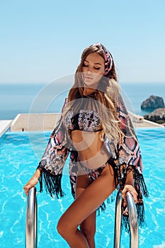 Beautiful woman with blond hair in elegant bikini posing near swimming pool with sea view