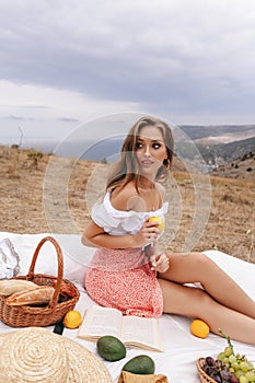 Beautiful woman with blond hair in elegant dress having picnic with mountains view