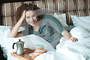 Beautiful woman with blonde curly hairstyle eat breakfast in bed with coffee and donuts