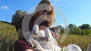 Beautiful woman with blonde hair sitting on grass at field and stroking her siberian husky dog. Young girl in sunglasses