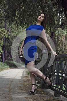 Beautiful woman in blue dress with red lips in a model pose on the sidewalk in the park is touching the parapet