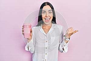 Beautiful woman with blue eyes drinking a take away cup of coffee pointing thumb up to the side smiling happy with open mouth