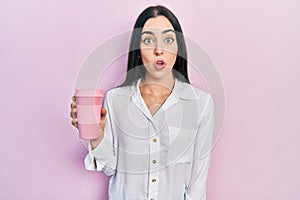 Beautiful woman with blue eyes drinking a take away cup of coffee scared and amazed with open mouth for surprise, disbelief face
