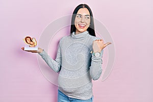 Beautiful woman with blue eyes expecting a baby, holding anatomic fetus pointing thumb up to the side smiling happy with open