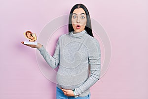 Beautiful woman with blue eyes expecting a baby, holding anatomic fetus scared and amazed with open mouth for surprise, disbelief