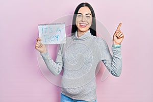 Beautiful woman with blue eyes expecting a baby holding its a boy banner surprised with an idea or question pointing finger with