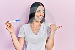 Beautiful woman with blue eyes holding pregnancy test result pointing thumb up to the side smiling happy with open mouth