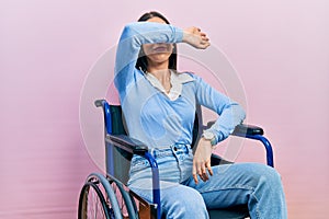 Beautiful woman with blue eyes sitting on wheelchair covering eyes with arm, looking serious and sad