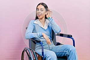 Beautiful woman with blue eyes sitting on wheelchair smiling with hand over ear listening an hearing to rumor or gossip