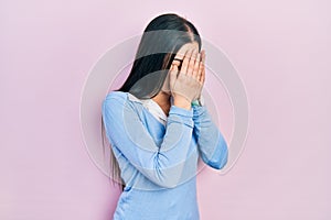 Beautiful woman with blue eyes standing over pink background with sad expression covering face with hands while crying