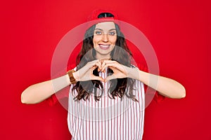 Beautiful woman with blue eyes wearing baseball sportswear and cap over red background smiling in love showing heart symbol and
