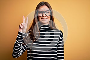 Beautiful woman with blue eyes wearing striped sweater and glasses over yellow background smiling with happy face winking at the