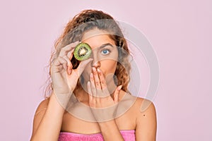 Beautiful woman with blue eyes wearing towel shower after bath holding kiwi fruit over eye cover mouth with hand shocked with