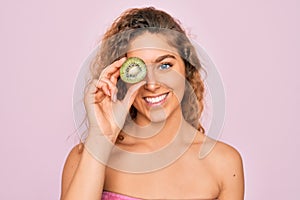 Beautiful woman with blue eyes wearing towel shower after bath holding kiwi fruit over eye with a happy face standing and smiling