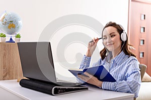 Beautiful woman in blue shirt with headphones and notebook in her hands sitting at laptop, online education