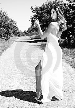 Beautiful woman, bride hitchhiker in white dress thumbs for a lift on a country road while showing her leg