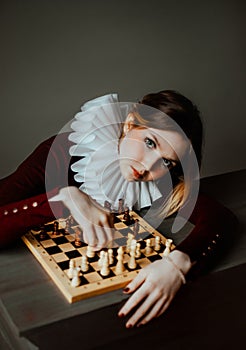 A beautiful woman in burgundy clothes and a white jabot sits at a table and plays chess. The Queen's gambit. Mind and logic.