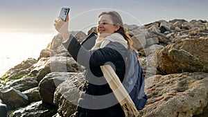 Beautiful woman capturing the essence of the stormy winter sea by taking pictures with her smartphone while standing on a rocky