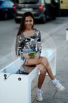 Beautiful woman charging her phone on free multipurpose solar panel charger incorporated in to sitting bench for citizens.Modern t