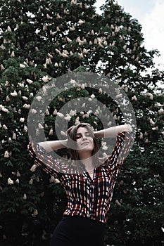 Beautiful woman in checkered shirt touches her hair and smiles to the camera near the blossomed tree