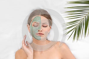 Beautiful woman with clay facial mask and tropical leaf on white fabric