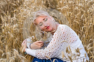 Beautiful woman with closed eyes sitting in golden wheat field and hug bunch of wheat ears. Liberty, love, happy summer concept