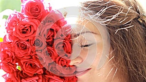 Beautiful woman closeup smiling and sniffing aroma of flowers of red roses. Wind in the hair girl.