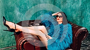 Beautiful woman in colorful green fur coat and big sunglases sitting in chair in high heel shoes. Fashion photo photo