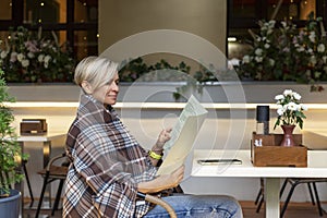 A beautiful woman in a cozy blanket at a table in a street cafe reads the menu. Relaxation in the city