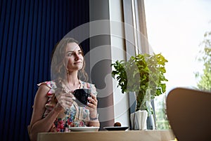 Young woman with cup of coffee in hands sitting at the table near a window in cafe. Coffee break time