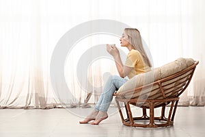 Beautiful woman with cup of hot drink relaxing in papasan chair at home, space for text