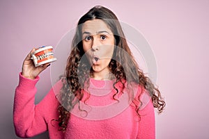 Beautiful woman with curly hair holding plastic teeth with dental braces over pink background scared in shock with a surprise