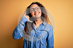 Beautiful woman with curly hair wearing casual denim shirt and glasses over yellow background smiling doing phone gesture with