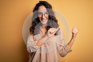 Beautiful woman with curly hair wearing striped shirt and glasses over yellow background Pointing to the back behind with hand and