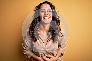 Beautiful woman with curly hair wearing striped shirt and glasses over yellow background smiling and laughing hard out loud