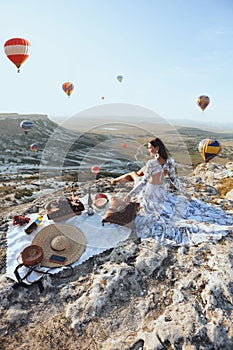 Beautiful woman with dark hair in elegant dress having picnic with fantastic view on valley with air balloons on background