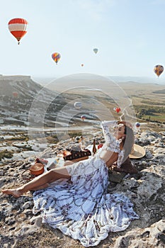 Beautiful woman with dark hair in elegant dress having picnic with fantastic view on valley with air balloons on background